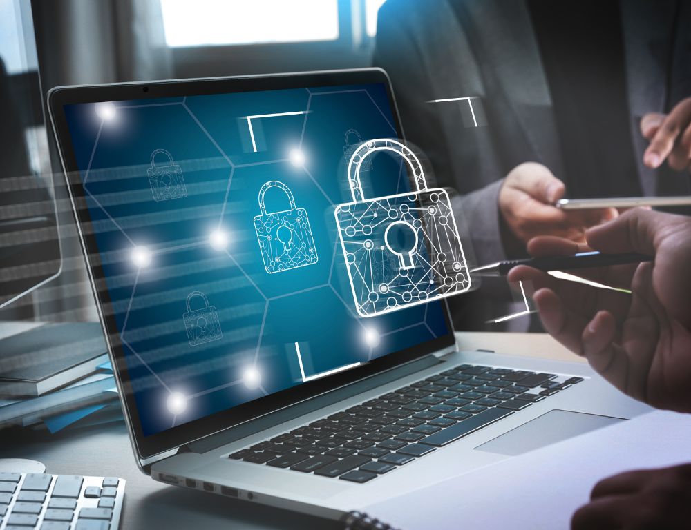 A laptop displaying a cybersecurity lock icon on its screen, with two people collaborating in the background.