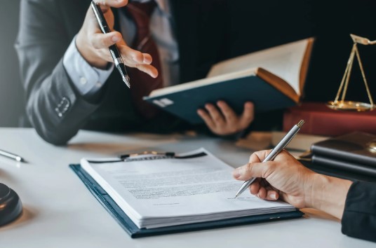 Two people in suits discuss a legal document at a desk.