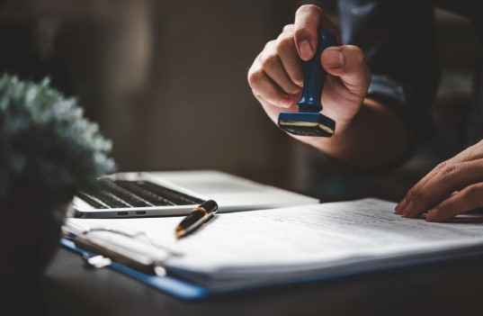 A person stamping a document on a desk with a laptop, pen, and paperwork.