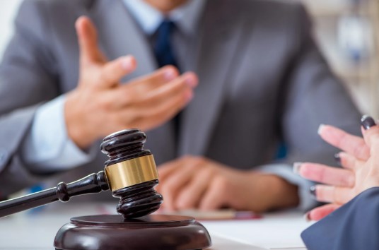 A close-up of two people in a formal setting, with one gesturing and a gavel on the table, suggesting a legal or business deal.