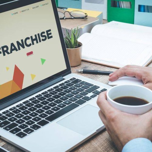 Hands holding a coffee mug over a laptop displaying the word "Franchise," with books and papers on a desk, illustrating franchise research and planning.